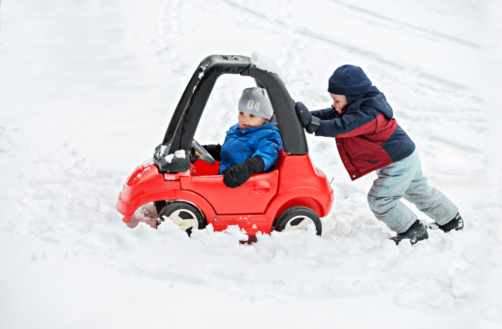 A young boy dressed for cold weather sits in a red toy car stuck in the snow during the winter season. His older brother helps by giving the car a push from behind.